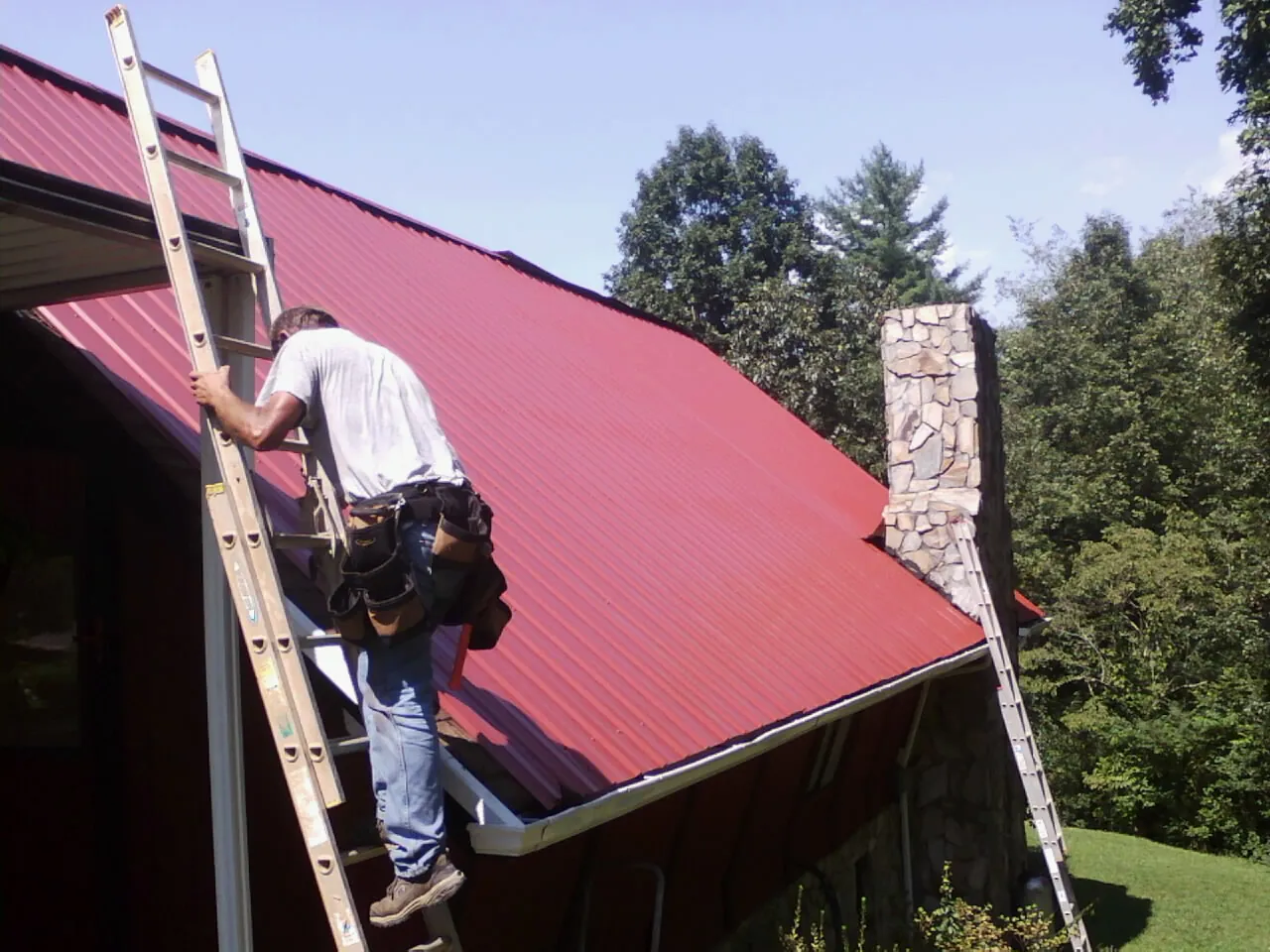 Roofer on a ladder beside a red metal roof and stone chimney — Soffit & Fascia Repair by Roofing Brothers in Dateland